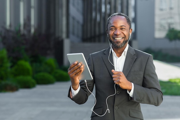 Exitoso jefe afroamericano fuera del edificio de oficinas con tableta y auriculares hablando