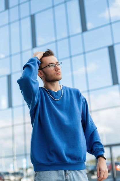 Exitoso joven apuesto hombre de negocios con gafas y cabello en un suéter azul de moda camina por la ciudad cerca de un edificio de vidrio de negocios