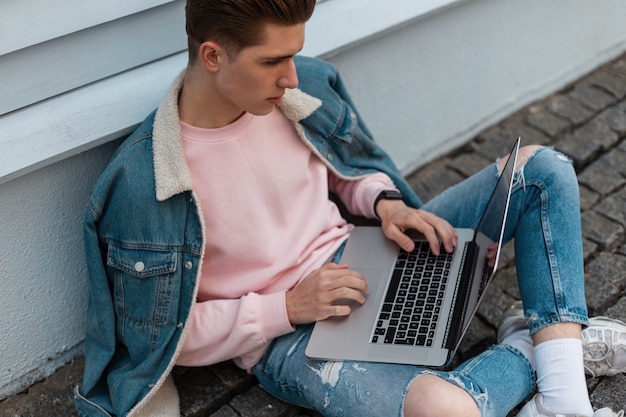 El exitoso joven independiente en ropa de jeans casual de mezclilla con estilo se sienta con la computadora portátil y trabaja de forma remota en un proyecto creativo. Diseñador de moda atractivo está escribiendo en el teclado. Retire el trabajo.