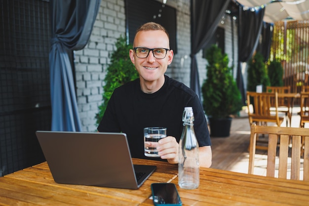 Un exitoso trabajador independiente con una camiseta negra y anteojos se sienta en la terraza de verano de un café y trabaja en una computadora portátil El gerente se sienta en un café durante el almuerzo y trabaja en línea