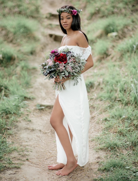 Exótica novia con piel oscura en vestido blanco con ricos bouquet rojo se encuentra en el parque