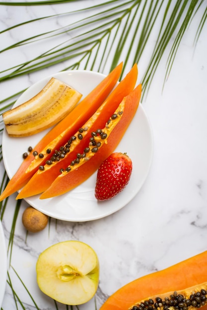 Exotisches Frühstück. Konzept für Sommer, Urlaub und gesunde Ernährung. Geschnittene Banane, Papaya, Erdbeere auf weißem Teller. Tropisches Blatt auf dem Tisch. Flache Lage, vertikaler Schuss.