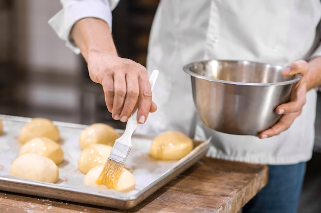 Experiencia, habilidad. Manos masculinas cubriendo hábilmente los rollos acostado sobre una bandeja para hornear en la mesa con glaseado de pastelería antes de hornear