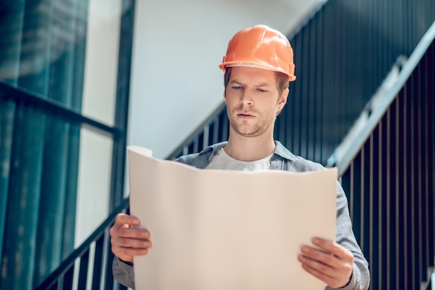 Experiencia laboral. Hombre concentrado en casco de seguridad naranja mirando de cerca el plan de construcción en papel de pie en el sitio de construcción