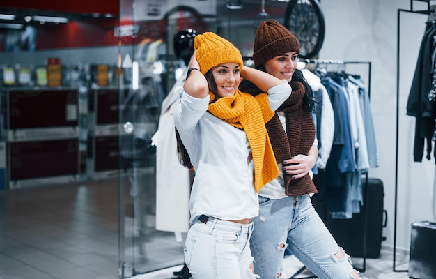 Experimentando chapéus e lenços quentes. Duas jovens fazem compras juntas no supermercado.