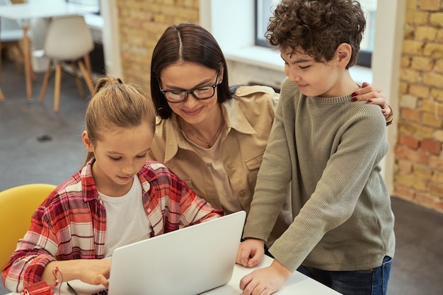 Experimentieren Sie mit einer fürsorglichen jungen Lehrerin mit Brille, die lächelt, während Sie intelligenten Kindern beim Lernen helfen