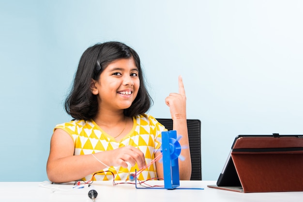 Experimento electrónico: pequeña estudiante india asiática que realiza una investigación de molinos de viento con cables, conexiones, estudia desde una computadora portátil o tableta