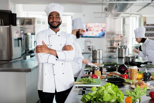 Experto en gastronomía de pie en la cocina profesional del restaurante con los brazos cruzados mientras sonríe a la cámara. Jefe de cocina confiado con uniforme de cocina mientras prepara los ingredientes para el servicio de cena.