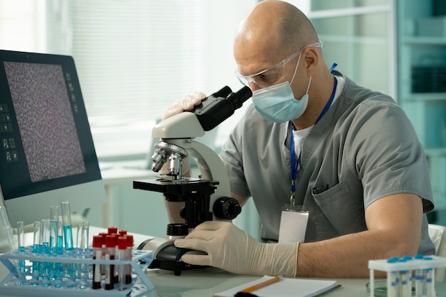 Experto de laboratorio concentrado en máscara y guantes sentado en un escritorio con gradilla para tubos de ensayo y examinando la muestra bajo el microscopio