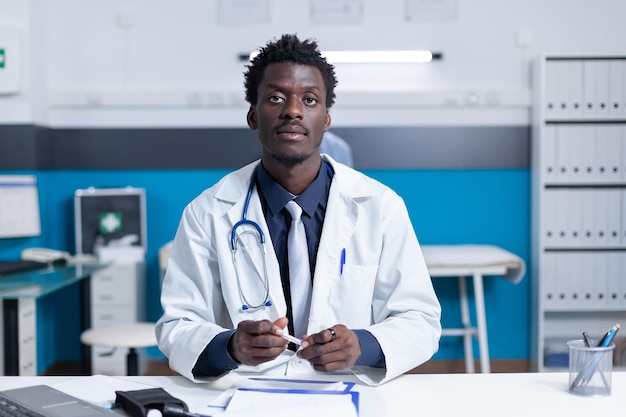 Experto en medicina general del policlínico afroamericano sentado en un gabinete clínico moderno con estetoscopio. Especialista médico del hospital en el trabajo sentado en el escritorio en el espacio de trabajo médico.