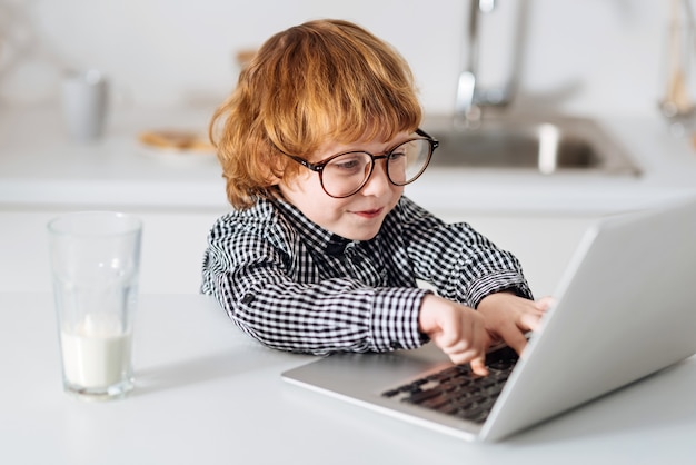 Explorando ideas. Divertido niño pelirrojo inteligente escribiendo una prueba en su computadora portátil mientras usa gafas y está sentado a la mesa en una cocina iluminada por el sol