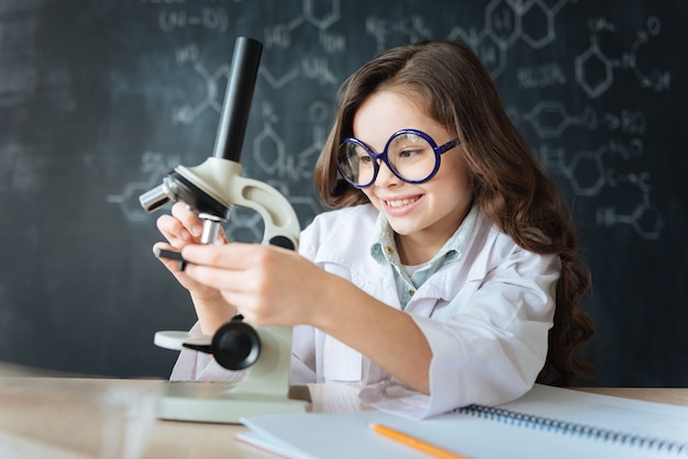 Explorando el mundo molecular. Investigador hábil y astuto sonriente sentado en el laboratorio y disfrutando de la clase de química mientras participa en el experimento de microbiología y usa microscopio