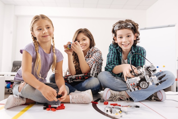 Explorando nuevos horizontes con amigos. Niños lindos, animados y sinceros, sentados en el aula de ciencias y jugando con aparatos y dispositivos mientras expresan felicidad