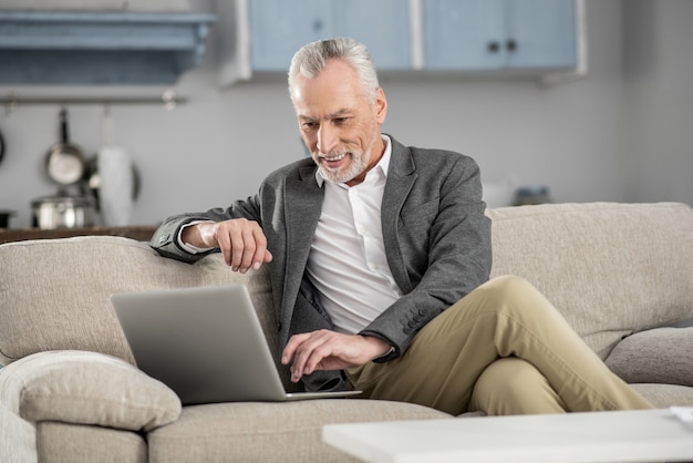 Expresa positividad. Hombre alegre manteniendo una sonrisa en su rostro y apoyado en el sofá mientras mira su computadora portátil