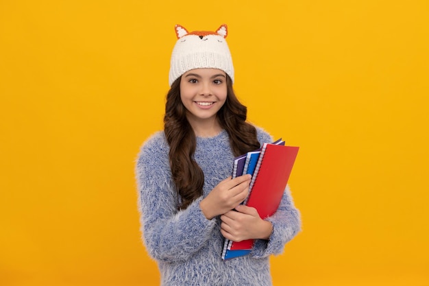 expresar emociones positivas. moda de invierno chico positivo con pelo rizado con sombrero. De vuelta a la escuela. jovencita en géneros de punto sobre fondo amarillo. retrato de niño con libro de trabajo.