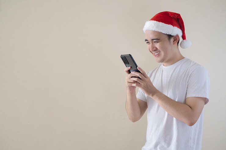 La expresión facial feliz del joven Papá Noel asiático mientras mira un teléfono inteligente en un fondo aislado