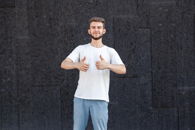 Expresión gestual y concepto de personas Hombre caucásico barbudo feliz de cabello oscuro con camiseta blanca y jeans mostrando los pulgares hacia arriba con la mano derecha en la calle sobre un fondo oscuro con espacio para copiar