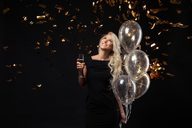 Foto expresiones brillantes de emociones felices de la increíble chica rubia celebrando la fiesta. vestidos negros de lujo, sonrientes, una copa de champagne, oropeles dorados, globos, cabello largo y rizado