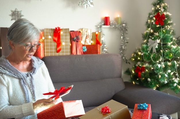 Expressão de surpresa de uma mulher idosa recebendo um novo tablet como presente de Natal. Cabelo grisalho e óculos. Muitos presentes ao redor dela