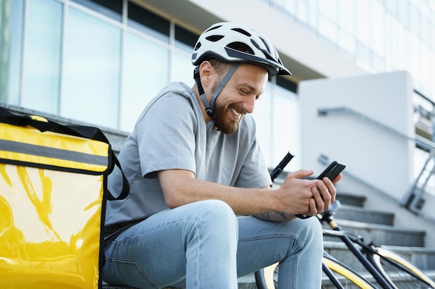 Expresskurier für die Lieferung von Lebensmitteln, der mit isolierter Tasche und Fahrrad auf der Treppe sitzt