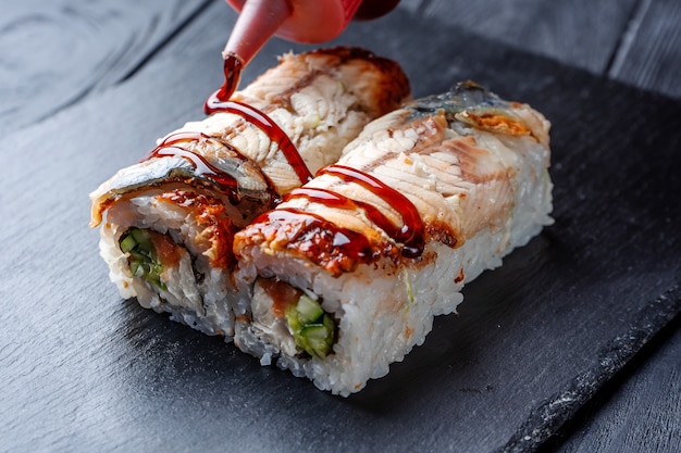 Exprima la salsa marrón en un conjunto de rollos de sushi con queso crema, arroz y salmón en un tablero negro decorado con jengibre y wassabi en una mesa de madera oscura. Comida japonesa. Enfoque suave selectivo