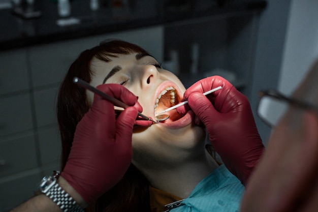 Extensivo examen dental. Ciérrese encima del tiro de una mujer joven que hace que sus dientes sean examinados por un dentista profesional del hombre que lleva guantes médicos rojos. Concepto oral dental