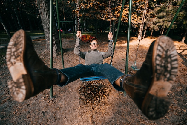 Foto extraño concepto de personas excéntricas extrañas. hombre adulto en botas de moda vintage montar swing en el parque de la ciudad en el patio de recreo para niños piernas abiertas