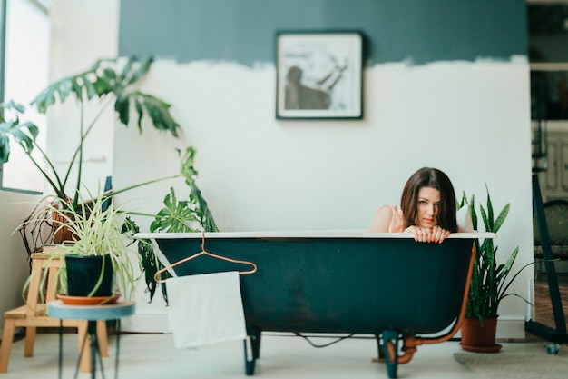 Extraño retrato de joven hermosa en vestido rosa acostado en el baño de hierro fundido vintage vacío interior decorativo en sala de estilo francés con plantas verdes.