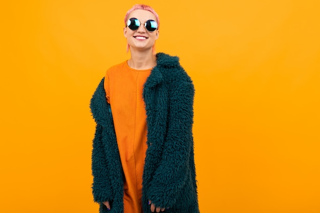 Extraordinaria mujer hermosa con cabello rosado corto en abrigo oscuro y gafas de sol sonrisas aisladas en naranja