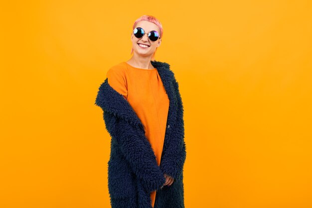 Extraordinaria mujer hermosa con el pelo corto de color rosa en abrigo oscuro y gafas de sol sonríe aislado sobre fondo naranja.