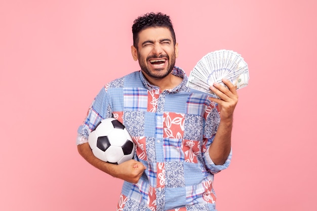 Extrem aufgeregter glücklicher Mann mit Bart in blauem Freizeithemd mit Fußball und Dollarnoten, die Wetten schreien und gewinnen, hält die Augen geschlossen Studioaufnahme im Innenbereich isoliert auf rosa Hintergrund