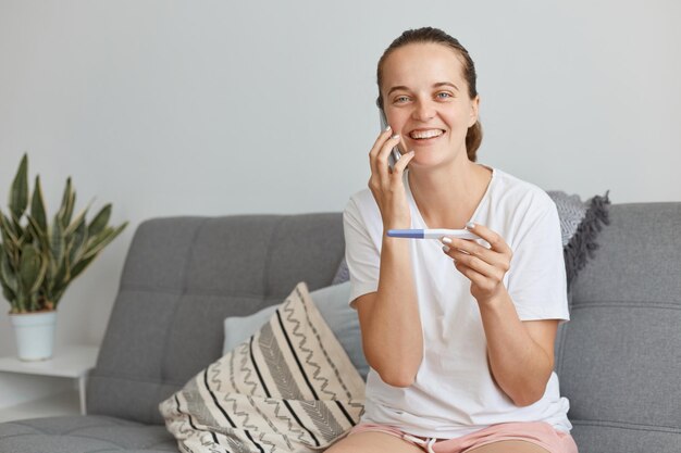 Extrem glückliche attraktive dunkelhaarige Frau mit weißem T-Shirt und kurzem, die auf dem Sofa im Wohnzimmer sitzt und telefoniert, Nachrichten über ihre Schwangerschaft mit positivem Test in den Händen erzählt.