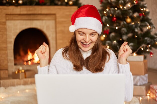 Extrem glückliche Frau, die am Laptop in der Nähe von Weihnachtsbaum und Kamin arbeitet, Weihnachtsmann-Hut und weißen Pullover trägt, Fäuste mit Erfolg ballt und im Wohnzimmer mit Weihnachtsdekorationen posiert.