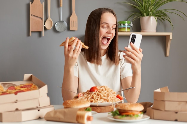 Extrem glückliche, überglückliche kaukasische Frau mit braunen Haaren und weißem T-Shirt, die in der Küche am Tisch sitzt und ein Stück Pizza in den Händen hält, die mit dem Handy mit Junk Food zu Abend essen