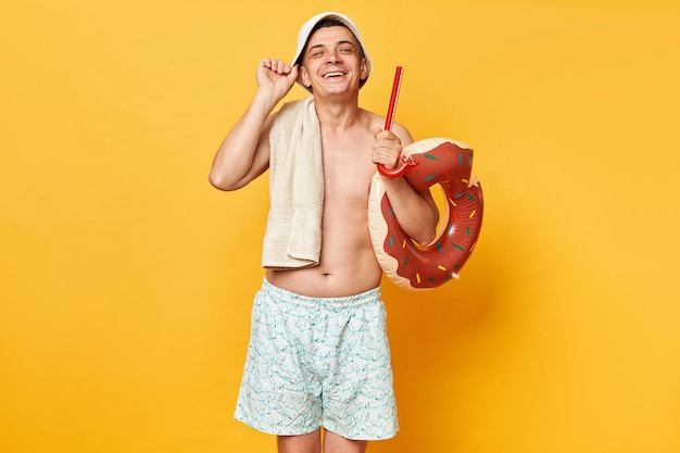 Extrem glücklicher, fröhlicher Mann in Shorts, Badeanzug und Panama hält einen Donut-Rubberring, isoliert auf gelbem Hintergrund und genießt einen ruhigen Sommerurlaub