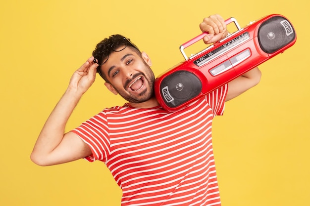 Extremadamente feliz hombre satisfecho con barba en camiseta roja a rayas sosteniendo una grabadora de cinta roja en los hombros, disfrutando escuchando música. Disparo de estudio interior aislado sobre fondo amarillo