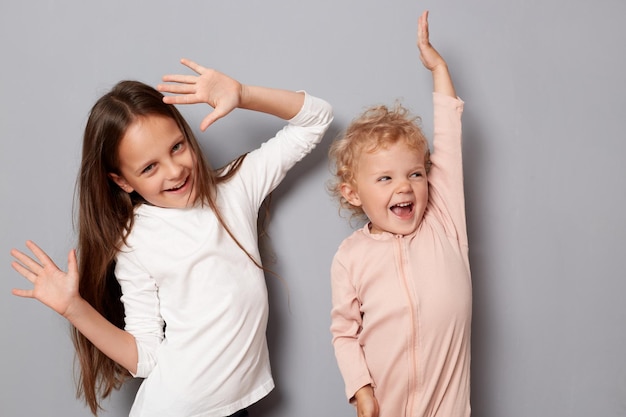 Extremamente felizes meninas menores irmãs se divertindo juntos dançando braços levantados brincando rindo com expressões alegres alegres de pé isolados sobre um fundo cinza