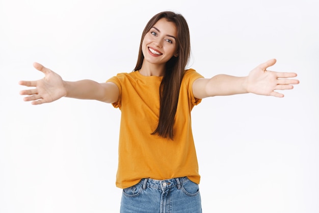 Foto extrovertida, feliz mujer morena agradable con camiseta amarilla, extendiendo las manos hacia los lados, inclinando la cabeza y sonriendo, queriendo que un amigo se acerque y dé un cálido abrazo, saludo a la amante, de pie, fondo blanco, abrazos