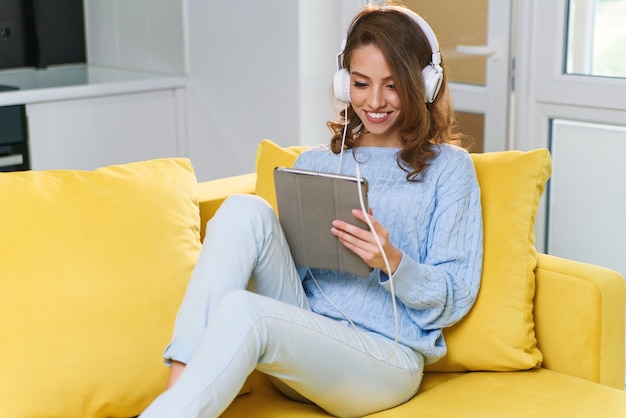 Exuberante garota de 30 anos, com cabelos castanhos ondulados, usando fones de ouvido brancos, ouve música e usa laptop ou tablet, descansando no sofá amarelo em um lar aconchegante.