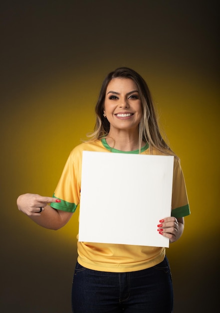 Fã brasileiro segurando um fã de mulher de cartão branco em branco comemorando no futebol ou partida de futebol em fundo amarelo cores do Brasil