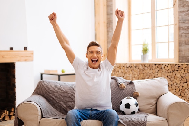 Foto fã de futebol. adolescente bonito e bem construído sorrindo e segurando os braços enquanto está sentado no sofá e com uma bola de futebol perto dele