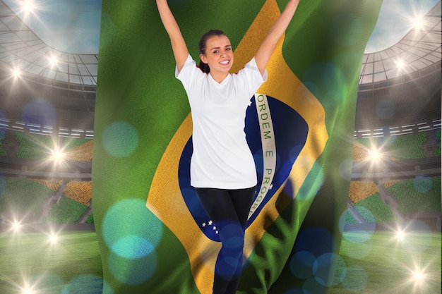 Foto fã de futebol animado em branco torcendo segurando a bandeira do brasil contra grande estádio de futebol com fãs brasileiros