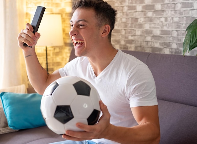 Fã de futebol animado menino assistindo a um jogo na televisão segurando uma bola de futebol na mão enquanto ele se senta em um sofá confortável em sua sala de estar