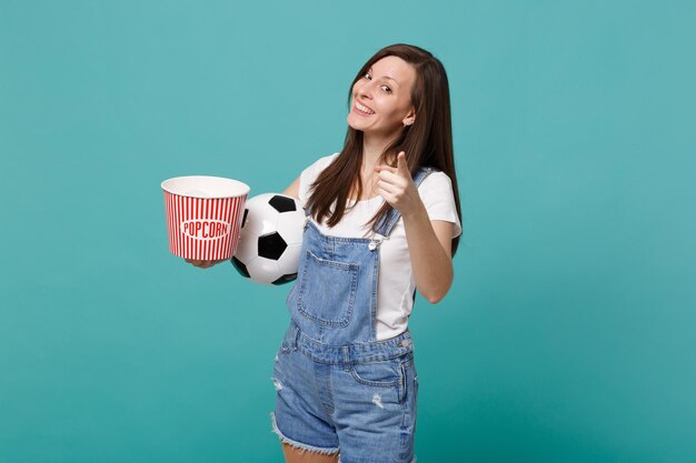 Fã de futebol de linda garota assistindo jogo de apoio ao time favorito com bola de futebol, balde de pipoca apontando o dedo na câmera isolada no fundo azul turquesa. Conceito de estilo de vida de lazer familiar esportivo.