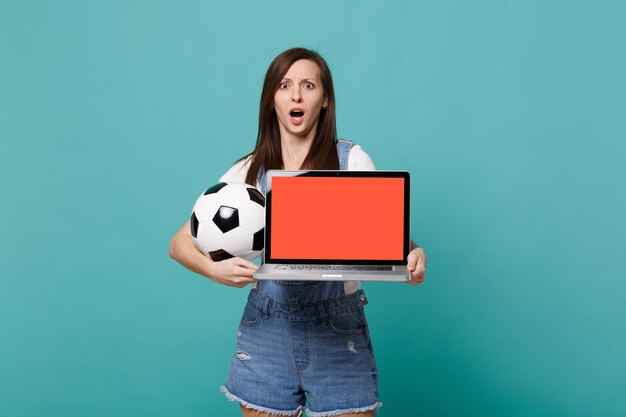 Fã de futebol de mulher chocada segurando o computador de computador portátil de bola de futebol com tela vazia em branco isolada no fundo da parede azul turquesa. Emoções de pessoas, conceito de lazer familiar esportivo. Mock up espaço de cópia.