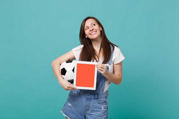 Fã de futebol de mulher sonhadora segurando uma bola de futebol, computador tablet pc com tela vazia em branco isolada em fundo azul turquesa. Emoções de pessoas, conceito de lazer familiar esportivo. Mock up espaço de cópia.