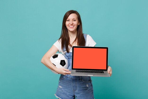 Fã de futebol de mulher sorridente segurando computador de pc portátil de bola de futebol com tela vazia em branco isolada no fundo da parede azul turquesa. Emoções de pessoas, conceito de lazer familiar esportivo. Mock up espaço de cópia.