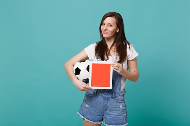 Fã de futebol de mulher sorridente segurando uma bola de futebol, computador tablet pc com tela vazia em branco isolada em fundo azul turquesa. Emoções de pessoas, conceito de lazer familiar esportivo. Mock up espaço de cópia.