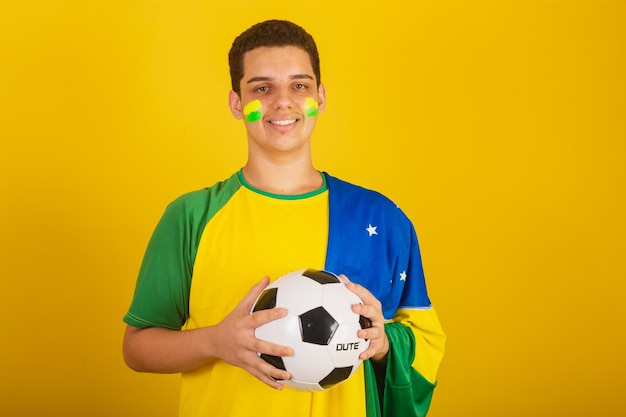 Fã de futebol jovem brasileiro vestido de verde segurando uma bola de futebol com bandeira do brasil