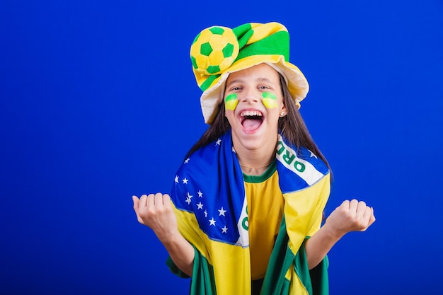 Fã de futebol jovem do brasil vestida de chapéu e bandeira usando espuma de dedo festejando incrível